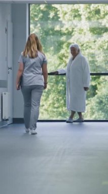 Hastane koridoru: Doktorlar ve profesyonel sağlık görevlileri yürüyor. Dijital tabletli hemşire pencerenin yanında duran yaşlı kadın hastaya geliyor. Klinikteki veya tıp merkezi koridorundaki tıbbi personel ve hastalar.