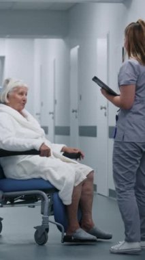 Erkek hemşire, kadın hastayla birlikte klinik koridorunda tekerlekli sandalye itiyor. Bayan doktor iş arkadaşına ve yaşlı hastasına gelir, konuşur, dijital tablet kullanır. Hastane koridorunda tıbbi personel ve hasta var..