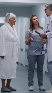Hemşire ve yaşlı kadın prosedürlerden sonra klinik koridorunda yürüyorlar. Erkek doktor iş arkadaşı ve hastasına gelir, test sonuçlarını dijital tablet kullanarak gösterir. Hastane koridorunda tıbbi personel ve hasta var..