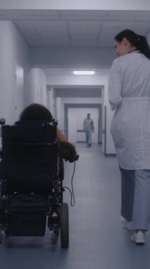 Kadın doktor klinik koridorunda yürüyor, fiziksel engelli bir kadına danışıyor. Doktorlar tekerlekli sandalyedeki hastalara sağlık kontrolünden bahsediyorlar. Modern tıp merkezinin koridoru. Dikey çekim
