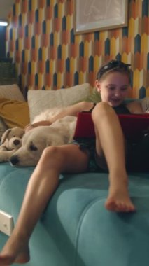 Genç kız kanepede köpekle oturuyor, tablette film izliyor, yavru köpeğe sarılıyor, gülümsüyor ve eğleniyor, evde boş zaman geçiriyor. Dikey çekim. Telefon ekranı görüntüsü. Golden retriever.
