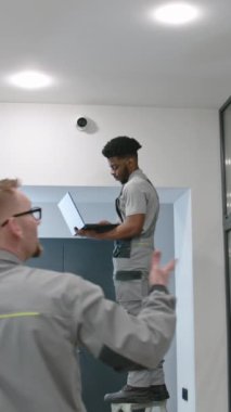 Two multi ethnic installers in uniform sets up security cameras in office corridor using laptop. Tired African American worker looks at camera. Concept of surveillance systems and privacy. Quick zoom.