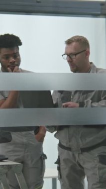 Two multi ethnic installers in uniform sets up security cameras or Internet in office room using laptop. They talk, check CCTV cameras in computer program. Concept of surveillance system and privacy.