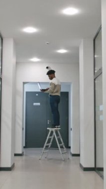 Afrikalı Amerikalı yükleyici, ofis odasına dizüstü bilgisayar kullanarak güvenlik kameraları kuruyor. Adam merdivende kalıyor ve bilgisayar programındaki güvenlik kameralarını kontrol ediyor. Gözetim sistemi ve mahremiyet kavramı. Dikey