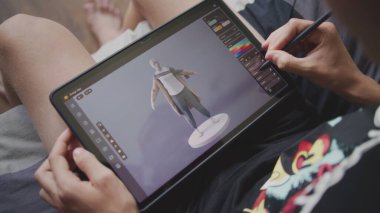 Odaklanmış genç, 3D modelleme programı kullanarak oyun karakteri için kıyafet çiziyor. Elinde tablet ve dijital kalemle odada oturan çocuk 3 boyutlu modeller yaratıyor. Oyun tasarımı ve geliştirme konsepti. Kapat..
