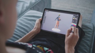 Odaklanmış genç, 3D modelleme programı kullanarak oyun karakteri için kıyafet çiziyor. Elinde tablet ve dijital kalemle odada oturan çocuk 3 boyutlu modeller yaratıyor. Oyun tasarımı ve geliştirme konsepti. Kapat..