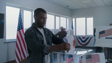 Afro-Amerikalı adam oy pusulasını doldurmak için oy kabinine giriyor. Çeşitli Amerikan vatandaşları ve seçmenler sandık merkezinde oy kullanıyor. ABD başkanlık seçimleri. Seçim kapsamı. Ulusun durumu.