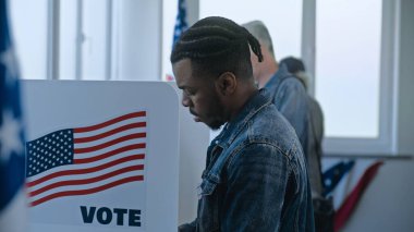Afro-Amerikalı adam seçim merkezindeki oy verme kabininde duruyor ve karar veriyor. Birleşik Devletler 'de Ulusal Seçim Günü. ABD başkan adaylarının siyasi ırkları. Vatanseverlik ve vatandaşlık görevi.
