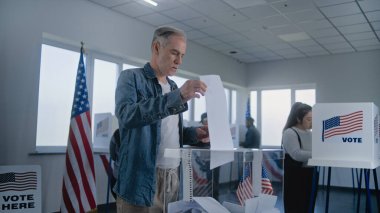 Yetişkin beyaz adam ilan kağıdını kutuya koyar. Çeşitli Amerikan seçmenleri ve Amerikan halkı oy kullanma merkezindeki oy kullanma kabinlerinde oy kullanıyor. Birleşik Devletler 'de başkanlık seçimleri. Vatandaşlık görevi. Yavaş çekim.