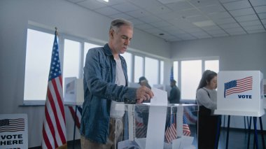 Yetişkin beyaz adam ilan kağıdını kutuya koyar. Çeşitli Amerikan seçmenleri ve Amerikan halkı oy kullanma merkezindeki oy kullanma kabinlerinde oy kullanıyor. Birleşik Devletler 'de başkanlık seçimleri. Vatandaşlık görevi. Yavaş çekim.