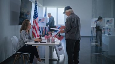 Beyaz, olgun bir Amerikan vatandaşı sandık merkezinde kayıt masasının yanında duruyor. Kadın anket memuru adamla konuşuyor, tablet bilgisayar kullanıyor ve oy vermek için oy pusulası veriyor. ABD 'de Seçim Günü.