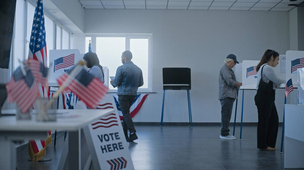 Афроамериканец берет бюллетень для голосования от женщины-участницы голосования за регистрационным столом и идет в кабинку для голосования. Многонациональные граждане США голосуют на избирательном участке. День выборов в США.