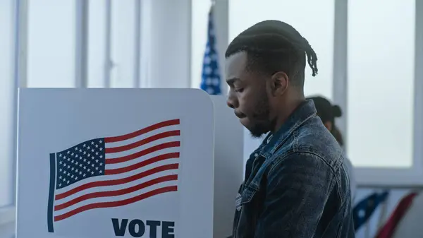 stock image African American man stands and decides at voting booth in polling station office. National Election Day in the United States. Political races of US presidential candidates. Patriotism and civic duty.