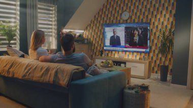 Beyaz çift televizyon ekranında Birleşik Devletler Seçim Günü 'nü izleyip evdeki kanepede oturup çay içiyorlar. Televizyonda yayınlanan son dakika haberiyle siyaset kanalı.