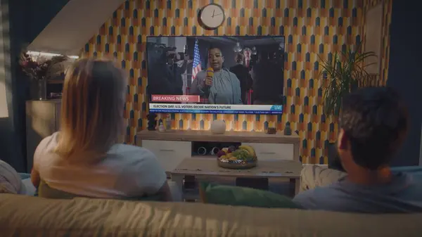 stock image Adult couple watches news broadcast report about United States elections on big TV screen sitting on sofa. TV display shows playback of breaking news with female African American reporter. Back view.