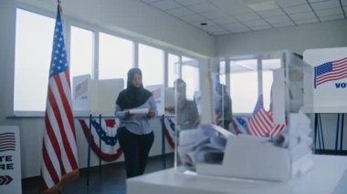 Başörtülü Müslüman kadın oy pusulasını kutuya koydu. Çok kültürlü seçmenler ve ABD vatandaşları sandık merkezindeki oy kullanma kabinlerinde oy kullanıyorlar. Birleşik Devletler 'de Ulusal Seçim Günü. Demokrasi kavramı. Yavaş çekim.