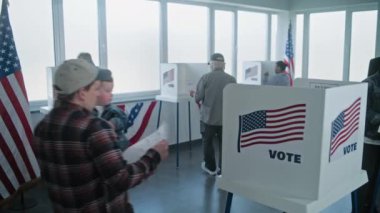 Küçük çocuklu beyaz bir adam oy pusulasını doldurmak için oy kabinine girer. Çok ırklı Amerikan vatandaşları ve seçmenler sandık merkezinde oy kullanıyor. ABD başkanlık seçimleri. Seçim kapsamı. Yurttaşlık görevi.