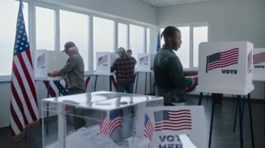 Afro-Amerikalı adam oy pusulasını doldurmak için oy kabinine giriyor. Çeşitli Amerikan vatandaşları ve seçmenler sandık merkezinde oy kullanıyor. ABD başkanlık seçimleri. Seçim kapsamı. Ulusun durumu.