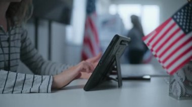 Kadın anket memuru oy kullanma merkezindeki kayıt masasında oturuyor, tablet bilgisayar kullanıyor ve oy pusulasını kişiye veriyor. Amerikan bayrağı masada duruyor. ABD başkanlık seçimleri. Kapat..