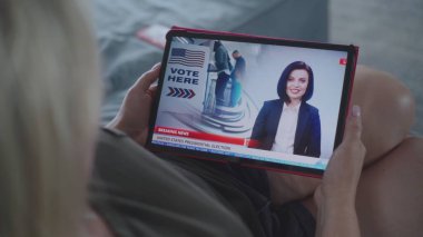 Tablet ekranda kadın haber spikeriyle televizyon haberleri yayınlanıyor. Kanepede yatarken dijital tablet kullanarak ABD başkanlık seçimlerini izleyen beyaz kadın. Kapat..