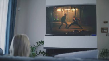 Yetişkin bir kadın rahat bir odada bir fincan kahveyle kanepede oturur ve televizyonda aksiyon filmi izler. Beyaz kadın hafta sonunu evde geçirir. Duvarda düz ekran TV monitörü ve gösterilen dövüş gerilim filmi.