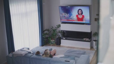 Yetişkin bir kadın modern oturma odasındaki kanepede dinleniyor ve ABD seçimleri hakkında TV haberleri izliyor. Genç kız odaya girer ve kanepeye oturur. Canlı yayın yapan büyük bir TV ekranı. Yüksek açı.