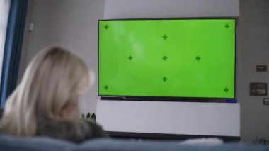 Yetişkin bir kadın rahat, modern bir oturma odasında bir fincan kahveyle kanepede oturur ve televizyon izler. Beyaz kadın dinleniyor ve hafta sonunu evde geçiriyor. Duvarında renkli anahtar yeşil ekran olan büyük bir televizyon..