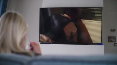 Yetişkin bir kadın rahat, modern bir oturma odasında bir fincan kahveyle kanepede oturur ve televizyon izler. Beyaz kadın dinleniyor, hafta sonunu evde geçiriyor. Duvarında aksiyon filmi olan büyük bir düz ekran TV monitörü.