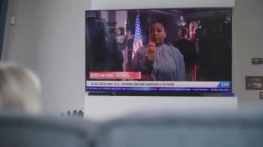 Kadın oturma odasında kanepede oturuyor ve televizyonda canlı yayın izliyor. Afrikalı Amerikalı kadın muhabir, ABD Seçim Günü 'nde canlı yayında son dakika haberlerini bildiriyor. Vatandaşlık görevi ve vatanseverlik kavramı.