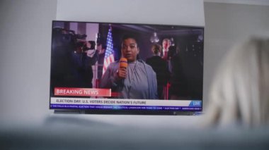 Kadın oturma odasında kanepede oturuyor ve televizyonda canlı yayın izliyor. Afrikalı Amerikalı kadın muhabir, ABD Seçim Günü 'nde canlı yayında son dakika haberlerini bildiriyor. Vatandaşlık görevi ve vatanseverlik. Arka plan.