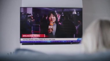 Kadın oturma odasındaki kanepede dinleniyor ve televizyonda canlı yayın izliyor. Asyalı kadın haber spikeri, Birleşik Devletler Seçim Günü 'nde canlı yayında son dakika haberlerini bildiriyor. Vatandaşlık görevi ve vatanseverlik. Arka plan.