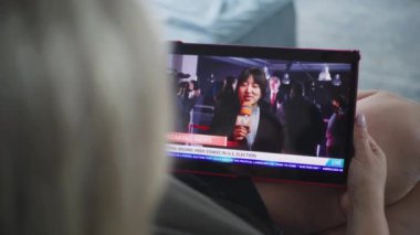 Kadın Asyalı muhabirin tablet ekranda yayınladığı son dakika gelişmeleri. Beyaz kadın, kanepenin üzerinde yatarken ABD seçimlerinin dijital tabletle yapıldığı haber bültenini izliyor. Kapat..