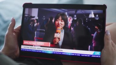 Tablet ekranında Asyalı kadın muhabirin son dakika haberlerinin görüntüleri var. Beyaz kadın, ABD seçimleriyle ilgili haber bültenini kanepede yatarken tablet bilgisayar kullanarak izliyor. Kapat..