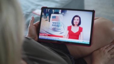 Kanepede uzanan tablet bilgisayarla Amerika Birleşik Devletleri başkanlık seçimleriyle ilgili son dakika haberlerini izleyen Kafkasyalı kadının yakın çekimi. Tablet ekranda kadın haber spikeriyle televizyon haberleri yayınlanıyor.