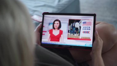 Beyaz bir kadının televizyon kanalında ABD seçimleri ve dijital tablet bilgisayarla oy kullanma ile ilgili haberleri izlerken kanepede uzanmasını kapatın. Tablet ekranda yayınlanan kadın haber spikeriyle flaş haber..