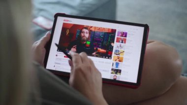 Kafkasyalı kadın, kanepede yatarken tablet bilgisayar kullanarak hisse senedi ticareti yapan blog yazarının eğitici videosunu izliyor. Tablet ekranda video içeriği ile video sunucu arayüzü gösteriliyor. Kapat..