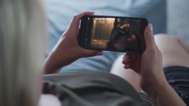 Yetişkin kadın koltukta uzanırken cep telefonu kullanarak aksiyon filmi izliyor. Gerilim filmli akıllı telefon ekranı. Beyaz kadın hafta sonları evde dinleniyor. Yaşam tarzı konsepti. Kapat..