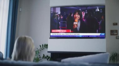 Yetişkin bir kadın, ABD seçimleri ve koltukta oturan büyük televizyon ekranında oylama ile ilgili haber bülteni izliyor. TV ekranında Asyalı kadın muhabirle son dakika haberlerinin görüntüleri var. Arka plan.