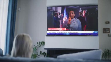 Yetişkin bir kadın, Birleşik Devletler seçimleriyle ilgili haber bültenini büyük TV ekranında, koltukta oturarak izliyor. TV şovu, Afrikalı Amerikalı kadın muhabirin son dakika haberlerini gösteriyor. Arka plan.