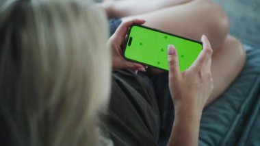 Elinde krom anahtarla cep telefonu tutan ve ekrana dokunan kadının arka görüntüsü. Beyaz kadın evdeki kanepede uzanıyor ve akıllı telefon kullanıyor. Yaşam tarzı konsepti. Yeşil ekran. Kapat..