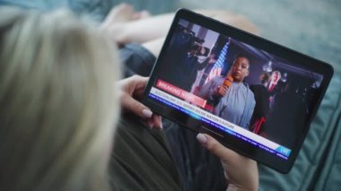 Afro-Amerikan kadın muhabirin tablet ekranda yayınlanan son dakika haberi. Beyaz kadın, kanepenin üzerinde yatarken ABD seçimlerinin dijital tabletle yapıldığı haberleri izliyor. Kapat..