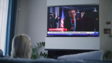 Yetişkin bir kadının, ABD Seçim Günü 'nü televizyon ekranında kanepede oturarak izlediği son dakika haberlerini izlemesi. Kadın gazetecinin TV ekranında görüntülendiği haber kaydı.
