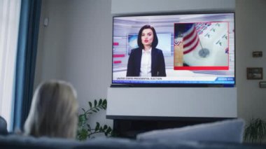 Yetişkin bir kadının, ABD başkanlık seçimlerini evdeki kanepede oturan büyük TV ekranında izlediği son dakika haberlerini izlemesi. TV 'de kadın haber spikeriyle televizyon haberleri yayınlanıyor.