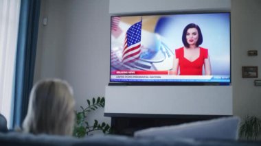 Yetişkin bir kadının, Birleşik Devletler seçimleri hakkındaki son dakika haberlerini izlediği büyük televizyon ekranında koltukta otururken. TV 'de haber sunucusunun görüntüleriyle haber bülteni oynatılıyor.