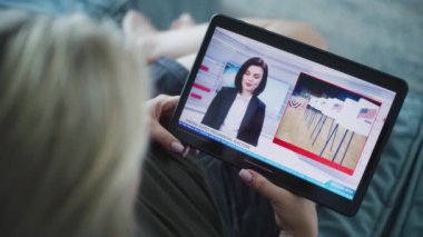 Tablet ekranda kadın haber spikeriyle televizyon haberleri yayınlanıyor. Beyaz kadın, kanepeye uzanmış dijital tablet kullanarak Birleşik Devletler başkanlık seçimleriyle ilgili son dakika haberlerini izliyor. Kapat..