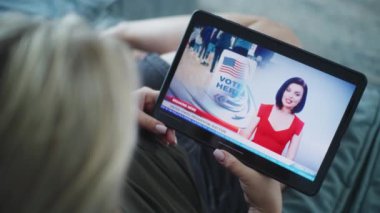 Kanepede yatarken tablet bilgisayar kullanarak ABD başkanlık seçimleriyle ilgili son dakika haberlerini izleyen beyaz kadının görüntülerini kapatıyoruz. Tablet ekranda kadın haber spikeriyle televizyon haberleri yayınlanıyor.