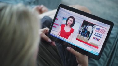 Beyaz bir kadının televizyon kanalında ABD seçimleri ve dijital tablet bilgisayarla oy kullanma ile ilgili haberleri izlerken kanepede uzanmasını kapatın. Tablet ekranda yayınlanan kadın haber spikeriyle flaş haber..