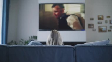 Şık bir oturma odasında rahat bir koltukta oturan ve evde dinlenirken TV 'de film izleyen bir kadının arkası. Büyük TV ekranında aksiyon filmi oynatılıyor. Ev sineması. Yaşam tarzı kavramı.