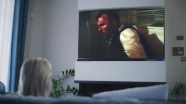 Televizyonda aksiyon filmi izleyen ve oturma odasındaki rahat koltukta oturan bir kadın. Yetişkin kadın hafta sonları evde dinlenir. Büyük TV ekranında film oynatılıyor. Ev sineması. Yaşam biçimi.