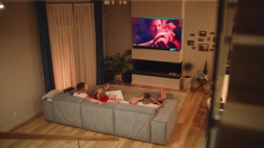 Kafkasyalı bir ailenin rahat bir oturma odasında kanepede uzanması ve büyük bir TV ekranında aksiyon filmi ya da TV şovu izlemesi. Ebeveynleri olan çocuklar akşamı evde geçirirler. Ev sineması. Yaşam tarzı kavramı.